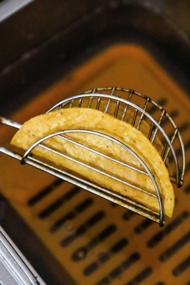 A taco fry holder with a crispy corn taco shell inside it held over a deep-fryer filled with hot oil.