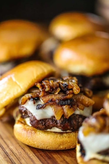 A close-up view of a slider that consists of a toasted slider bun with rosemary-garlic aioli, beef patty, melty provolone cheese, and topped with caramelized onions.