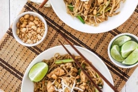 Two white bowls filled with chicken pad Thai with a small bowl of peanuts nearby.