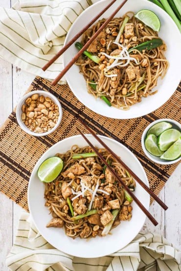 Two white bowls filled with chicken pad Thai with a small bowl of peanuts nearby.