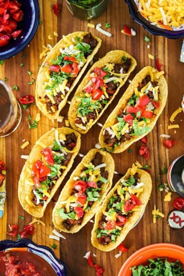 Six Tex-Mex Beef Tacos sitting up-right in two taco stands sitting on a cutting board surrounded by bowls of traditional toppings.