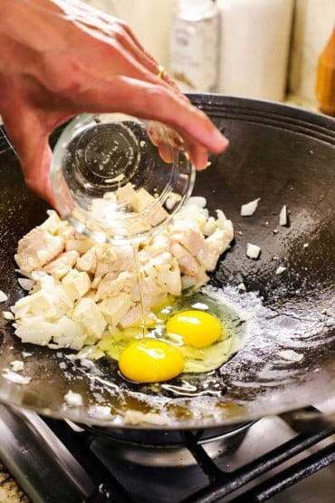 A person adding a couple of raw eggs into a wok that has cooked chicken pieces and onion pushed to one side.