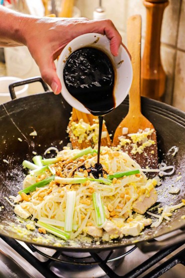 A person pouring a tamarind and soy sauce mixture into a wok filled with noodles, chicken, peanuts, and tofu.
