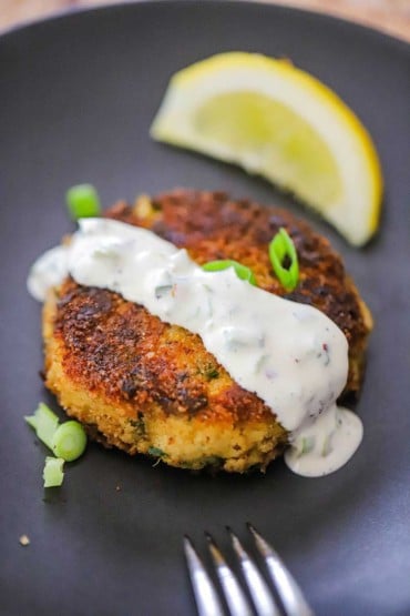 A close-up view of a Southern-style crab cake with a drizzle of jalepeño remoulade poured over the top and a lemon wedge next to it.