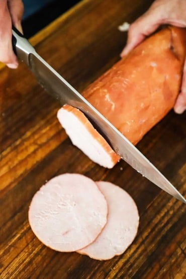 A person slicing discs of Canadian bacon from a large tube of the smoked meat on a cutting board with a chef's knife.