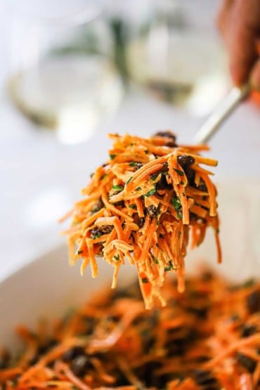 A person using a large serving spoon to lift a helping of carrot and raisin salad from a bowl.