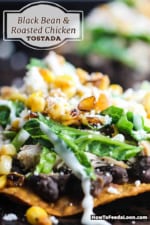 A close-up view of a black bean and roasted chicken tostada topped with shredded lettuce, corn salsa, Mexican crema, and crumbled Cotija cheese.