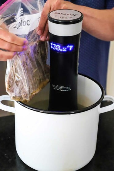 A person lowering an uncooked skirt steak in a plastic baggie into a pot of water with a sous vide machine inserted into it.