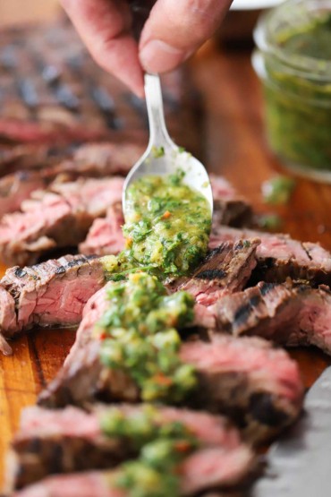 A person using a small spoon to spread chimichurri sauce over the top of sliced sous vide skirt steak.