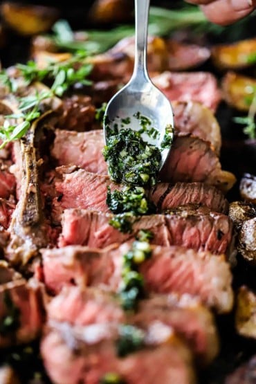 A person using a small silver spoon to drizzle sautéed herbs and garlic over slices of a medium-rare porterhouse steak.