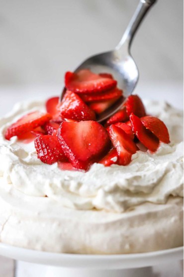 A close-up view of a silver spoon being used to place macerated strawberries on top of whipped cream that is resting on a layer of baked meringue.
