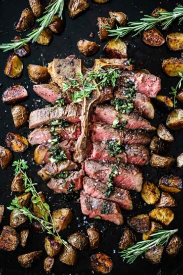 An overhead view of bistecca alla fiorentina that has been cut into slices, topped with sautéed herbs and surrounded by roasted balsamic potatoes.