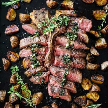 An overhead view of bistecca alla fiorentina that has been cut into slices, topped with sautéed herbs and surrounded by roasted balsamic potatoes.