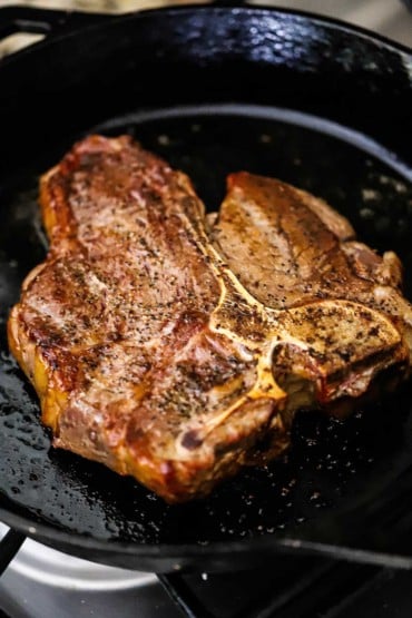 A close-up view of a porterhouse steak that is being seared in a hot cast-iron skillet.