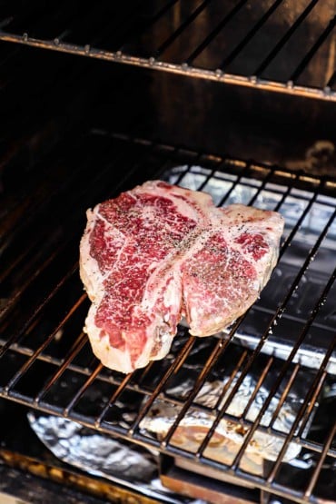 An uncooked porterhouse steak that has been seasoned with salt and pepper sitting on a grate inside an electric smoker.