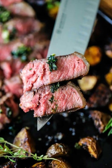 A large chef's knife holding up two slices of a bistecca alla Fiorentina with some sautéed herbs on each slice.