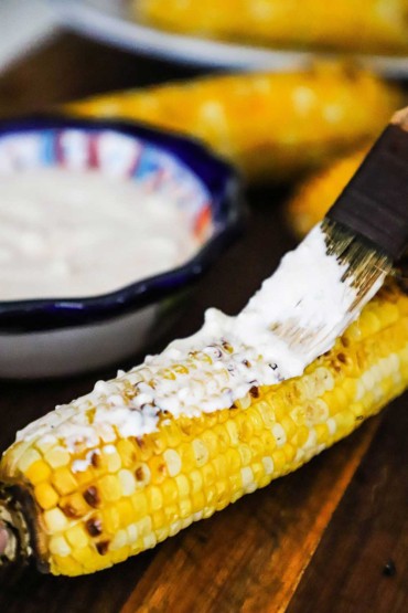 A person using a small brush to smear on a cream sauce all over a grilled ear of yellow corn.