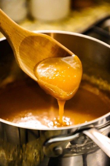 A wooden spoon that is being lifted from a saucepan of caramel and has caramel dripping from the end of the spoon.