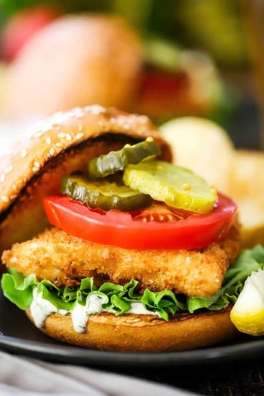 A close-up view of a fried fish sandwich that includes homemade tartar sauce, a slice tomatoes, green leaf lettuce, and bread and butter pickles on a toasted hamburger bun.