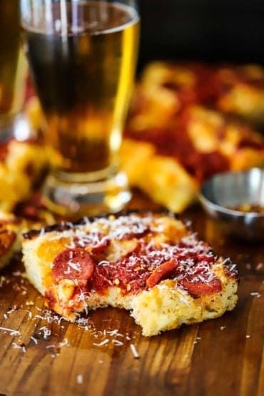 A square piece of Detroit-style pizza with a bite taken out of it and sitting next to a tall glass of beer.