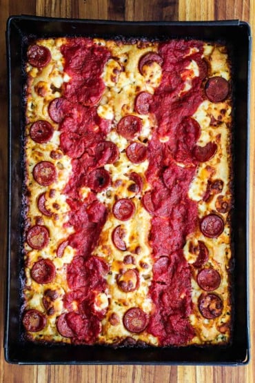 An overhead view of a whole Detroit-style pizza in a rectangular steel pan with two parallel stripes of red sauce on top of the pizza.