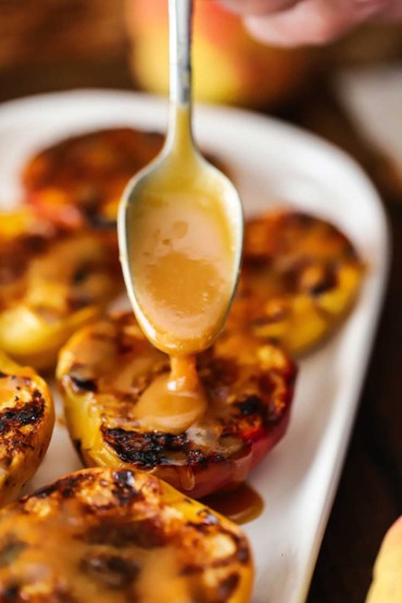 A person using a small spoon to drizzle homemade caramel sauce of a platter filled with grilled peaches.