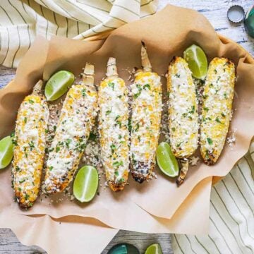 An overhead view of a row of elote on two sheets of brown wax paper and surrounded by wedges of lime.
