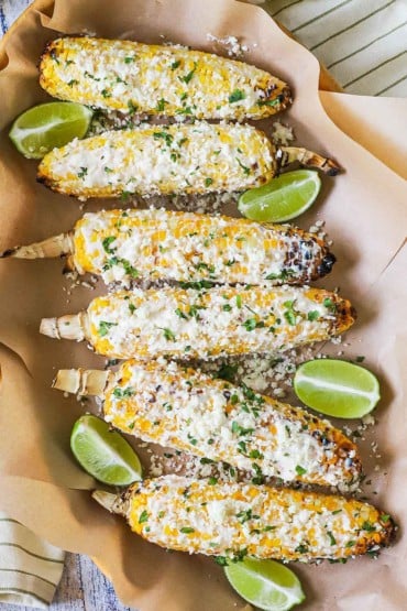 An overhead view of a row of elote on two sheets of brown wax paper and surrounded by wedges of lime.