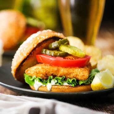 A close-up view of a fried fish sandwich that includes homemade tartar sauce, a slice tomatoes, green leaf lettuce, and bread and butter pickles on a toasted hamburger bun.
