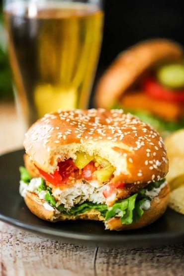 A straight-on view of a fried fish sandwich with a bite taken out of it sitting on a black dinner plate and a glass of beer in the background.
