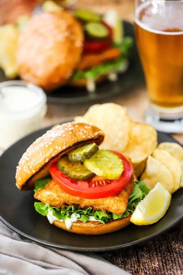 A fried fish sandwich sitting on a black plate next a lemon wedge and potato chips with a glass of beer and another plate of a fish sandwich in the background.
