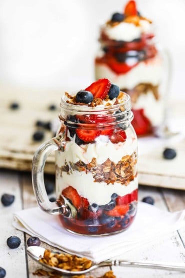 A straight-on view of a glass jar with a handle holding a wild berry parfait with mascarpone and topped with strawberries, blueberries, and granola.
