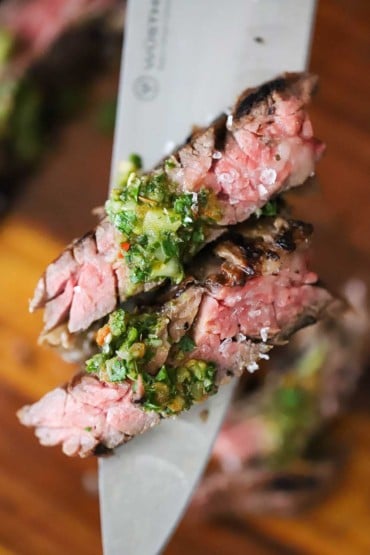 Two pieces of sous vide skirt steak sitting on a raised chef's knife and topped with a little chimichurri sauce and finishing salt.