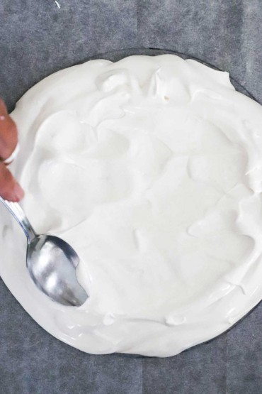 An overhead view of a person using a spoon to spread out French meringue into a circular mound on parchment paper.