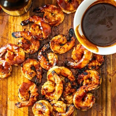 An overhead view of a pile of BBQ shrimp sitting on a cutting board with a small white bowl filled with homemade BBQ sauce nearby.