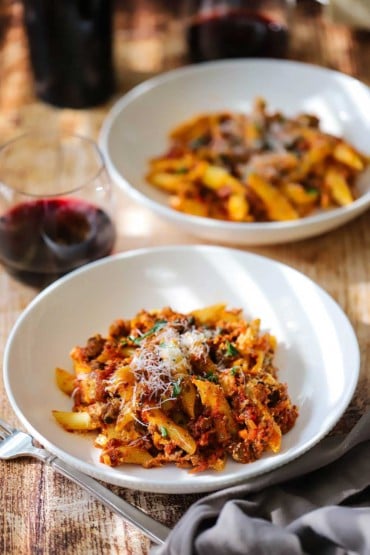 Two white pasta bowls filled with a dinner-sized portion of baked ziti topped with grated Parmesan cheese and sitting next to a stemless glass of red wine.