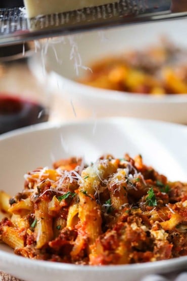 A close-up view of a white pasta bowl filled with baked ziti with a person grating a block of Parmesan cheese above it.