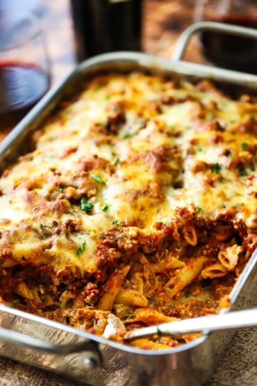 A straight-on view of a steel lasagna pan filled with baked ziti with about one third of the casserole gone and a large spoon inserted off to the side.
