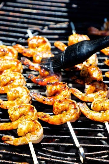 A person using a large rubber brush to apply barbeque sauce onto a row of shrimp that has been skewered and it on the grate of a charcoal grill.