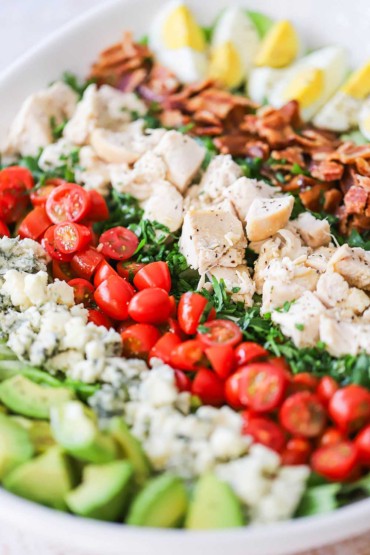 A close-up view of a Cobb salad that is lined with rows of chopped avocado, crumbled blue cheese, sliced tomatoes, cubed cooked chicken, crumbled bacon, and hard boiled egg wedges.