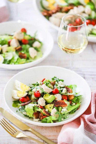 Two medium-sized white salad bowls filled with a tossed Cobb salad both sitting next to a glass of white wine.