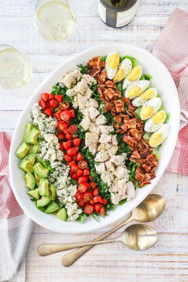 An overhead view of a Cobb salad on a large white oval platter flanked by two glasses of white wine and two gold serving spoons.