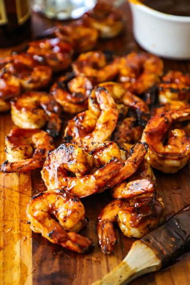 A straight-on view of a pile of grilled BBQ shrimp on a cutting board with a kitchen brush nearby that has been dipped in BBQ sauce.