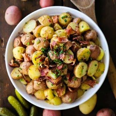 An overhead view of a white bowl holding a large serving of German potato salad with a few whole baby potatoes and petite dill pickles surrounding the bowl.