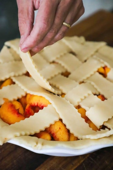 A person adding strips of homemade pie dough in a lattice formation over the top of an uncooked peach pie.