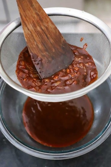 A person using a wooden spatula to press homemade BBQ sauce through a mesh sieve into a glass bowl, leaving sauteéd onions behind.