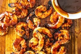 An overhead view of a pile of BBQ shrimp sitting on a cutting board with a small white bowl filled with homemade BBQ sauce nearby.