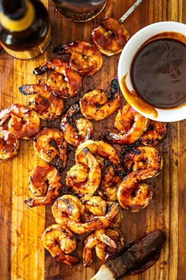 An overhead view of a pile of BBQ shrimp sitting on a cutting board with a small white bowl filled with homemade BBQ sauce nearby.