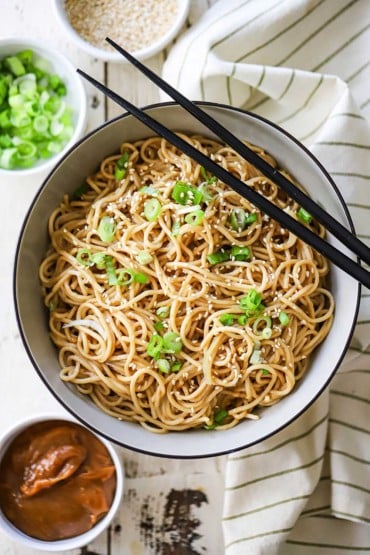 An overhead view of Chinese noodles with peanut sauce garnished with chopped scallions and sesame seeds.
