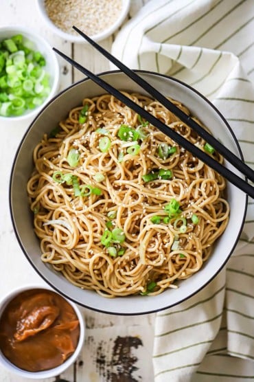An overhead view of Chinese noodles with peanut sauce garnished with chopped scallions and sesame seeds.